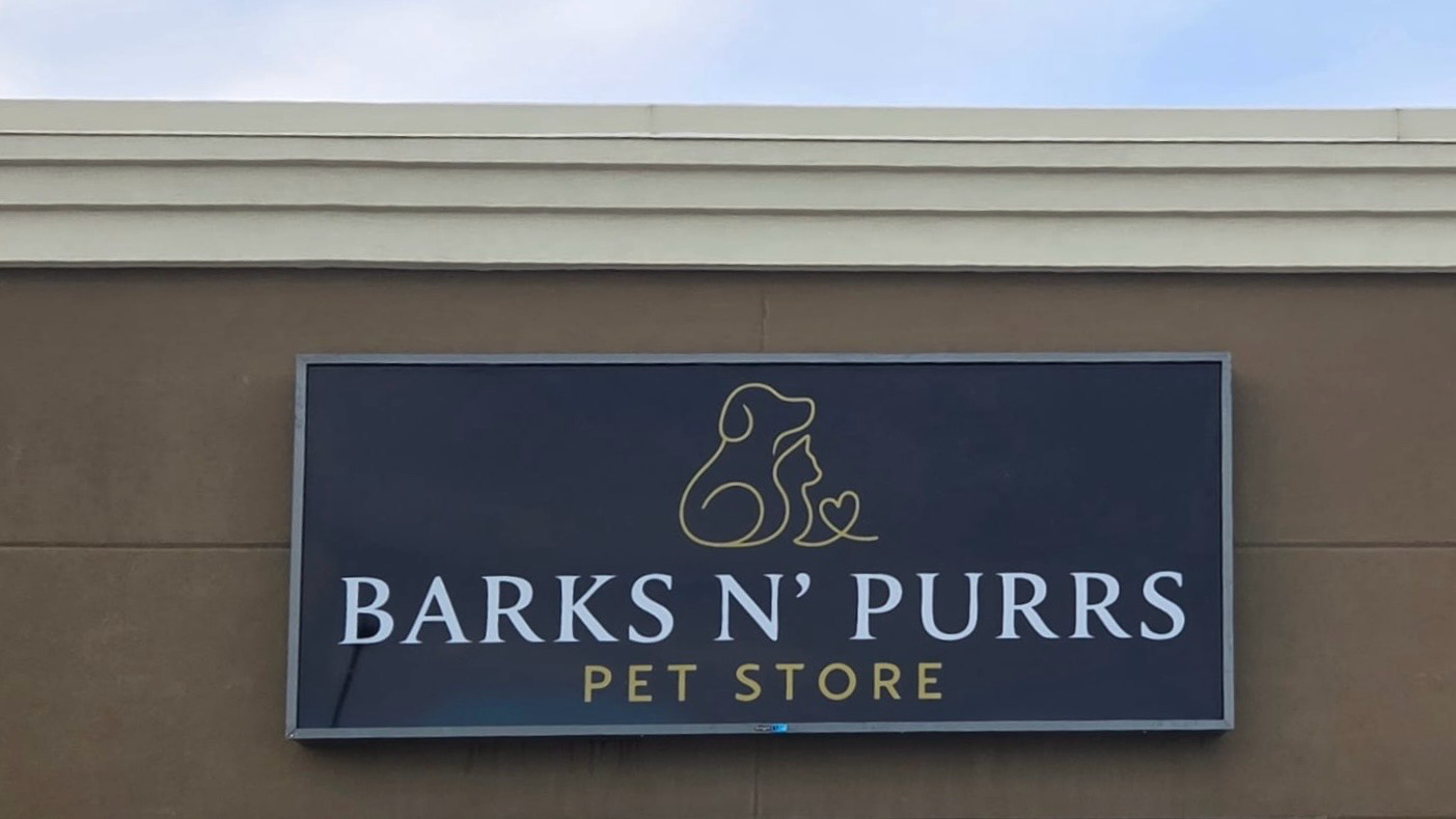 Photograph of the Barks N’ Purrs Pet Store storefront sign, with the dog and cat logo above white and gold lettering on a dark panel.